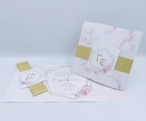 Προσκλητήρια γάμου πολυτελείας U011 Λευκό με ροζ απαλά λουλούδια, χρυσή λεπτομέρεια στη κοδελα και καρτάκι με μονογράμματα της εταιρίας  NewAge invitations